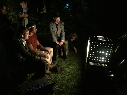Photographed from the side, two white men and a black man sit, a white man stands behind them and looks at his phone. The black man wears an EEG headset and looks at a glowing black box. The scene is outside on a grassy patch in front of a brush.