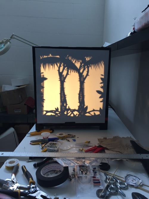 A black box with a scene depicted via silhouette: trees, flowers, vines. It sits on a white workbench with tools, tape, a ruler, and hardware all around it.