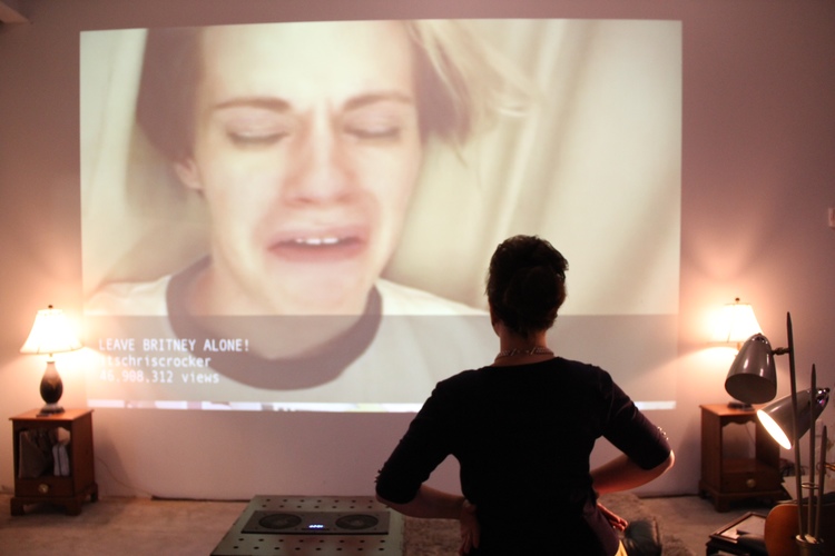 We see from behind: a white woman in a yellow dress and black cardigan stands, with her hands on her hip, next to a 20in black cube, projecting something onto the wall in the background. The projection is of the famous "Leave britney alone!" YouTube video. The scene is set like a living room, with side tables, lamps, and a rug on the floor.
