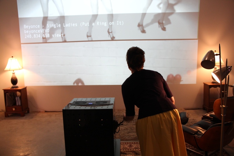 We see from behind: a white woman in a yellow dress and black cardigan leans over a 20in black cube, projecting something onto the wall in the background. She turns a metal crank on the side of the box.