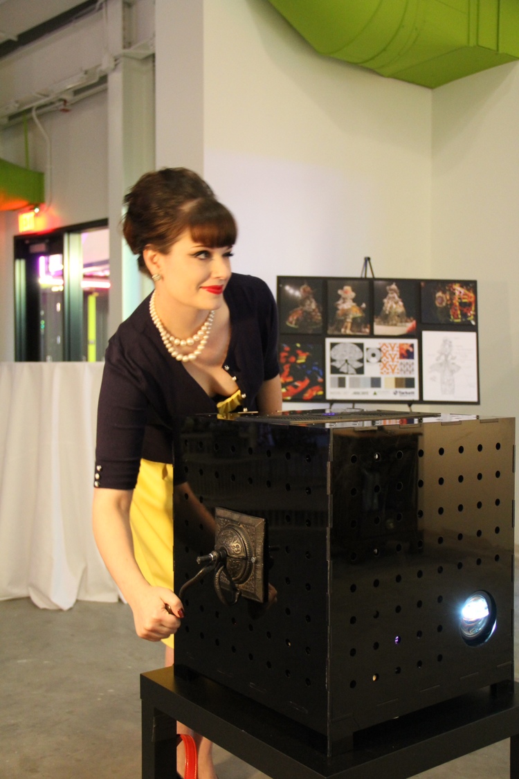 A white woman in a yellow dress and black cardigan leans over a 20in black cube with a projector bulb shining from it, projecting something out of frame. She turns a metal crank on the side of the box.