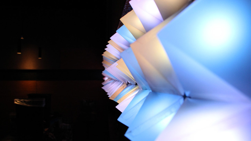 A sideview of the project: paper pyramids arranged next to one another in a 3 (high) by 48 (wide) array. They glow different shades of light blue, orange, and pink.
