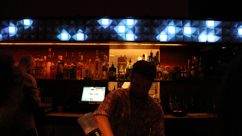 A white male bartender wearing a plaid shirt pours a drink in a dark bar. Behind him, an array of 3D pyramids (48 wide by 3 high) are randomly light up with white light. Most are dark.