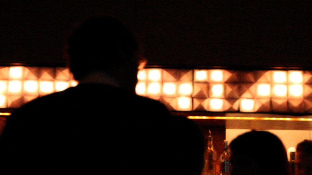 A man in a collared shirt stands with his back to camera, facing a dark bar. Behind the bar, an array of 3D pyramids (48 wide by 3 high) are randomly light up with orange light. Some are dark.