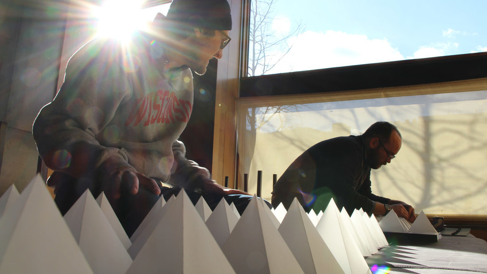 Two white men hunch over a bench with an array of paper pyramids. The sun shines behind them through windows of the room in which they work. The man in the forefront wears a beanie, has a hoodie that reads, "Wisconsin," and has a beard. The man in the back is not as legible.