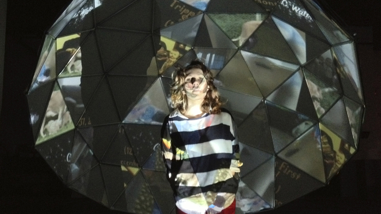 A white woman with a black and white thick horizontal striped shirt stands in front of a sculpture. The sculpture is a half-dome made of primarily triangular sections. Images are projected into each separate section. Because she stands between the projector and the sculpture, the images appear on her skin and clothes as well.