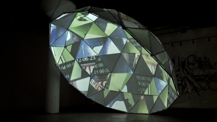 Wide shot of a sculpture—a half-dome made of primarily triangular sections. The sculpture is hanging from the ceiling on one side and touching the ground on the other. Images are projected into each separate section. The sculpture sits in a dark gallery with a white support column in the background.