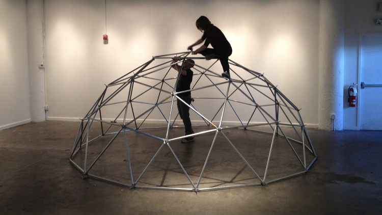 A large (14-foot wide) geodesic dome made of steel bars sits on the floor of an art gallery. Two people work on finishing its construction, one standing below the apex, one has climbed onto it. They are silhouetted.