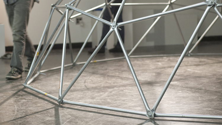 A tight shot of the triangular subsections of a geodesic dome made of steel beams. Someone in grey jeans, wearing grey sneakers, walks around it. 