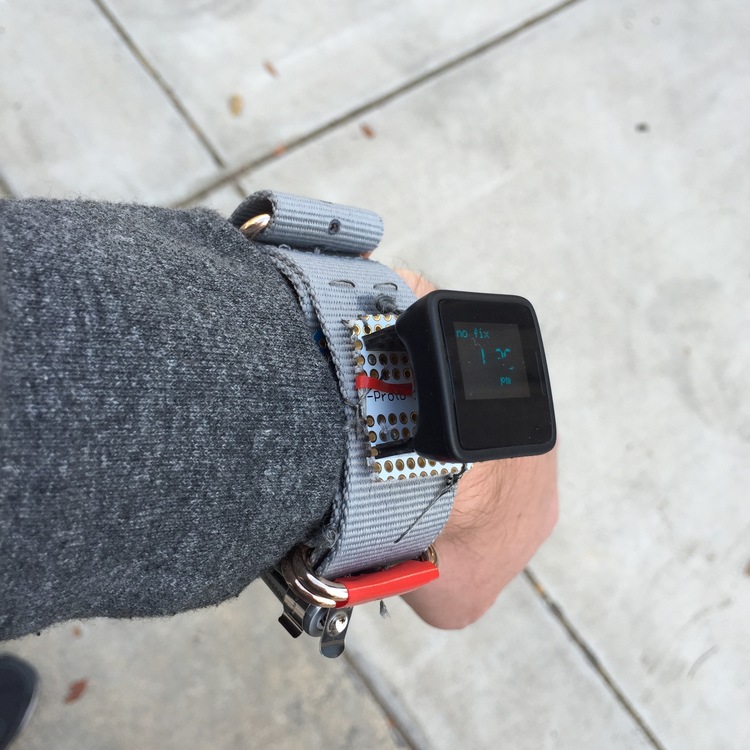 A white left arm with grey sweatshirt sleeve shot from the elbow down. On the wrist is a grey nylon watch strap. Instead of a traditional watch face, there is a small soldering board (1in square, white with a grid of small copper-rimmed holes) with a small black device attached. The device is 1in x 1in square and about .75in tall. It has a small LCD screen on it with green text on a black background. The text is not legible from this angle.