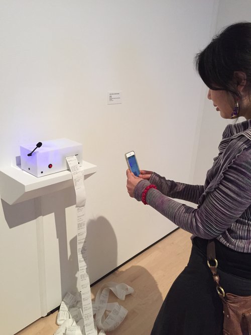 A white acrylic box with a small microphone coming out of the side sits on a shelf mounted on a white gallery wall. It glows a light blue. From the box, a long scroll of receipt paper flows. A woman with black hair is photographed from the side as she takes a photo of the box with her phone.